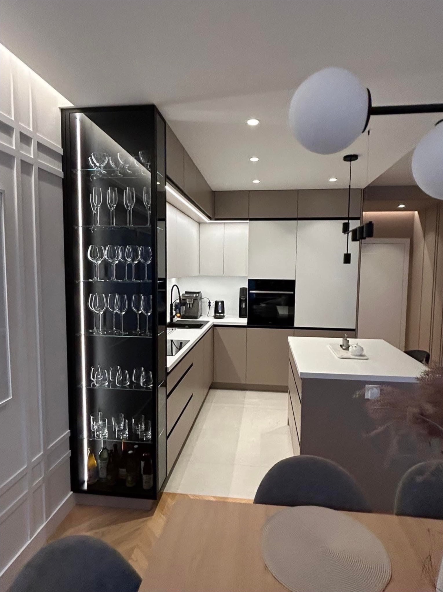 Nowoczesna kuchnia w beżach i bieli z wyspą, oświetleniem LED i przeszkloną witryną na alkohole. Minimalistyczny design z dbałością o detale.