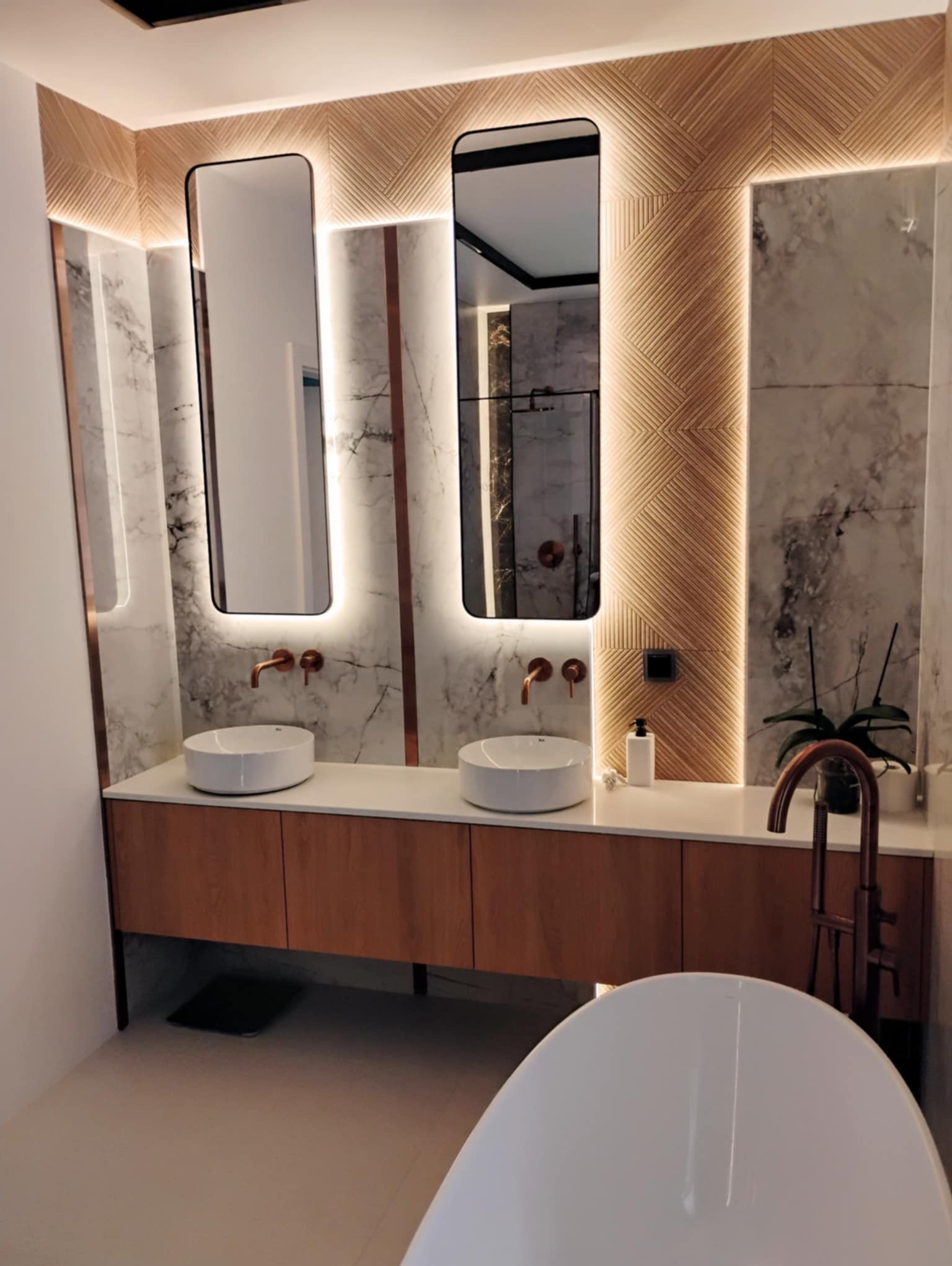 Nowoczesna łazienka z marmurowymi ścianami, drewnianą zabudową umywalkową i podświetlanymi lustrami w czarnych ramach. Widoczna wanna wolnostojąca i armatura w kolorze miedzi.