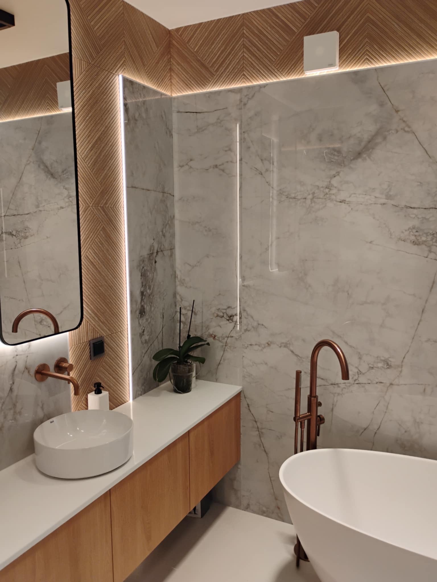 Elegancka łazienka w Chotomowie z marmurowymi płytkami, drewnianymi akcentami i miedzianą armaturą. Nowoczesny design z podświetlanym lustrem i wolnostojącą wanną.