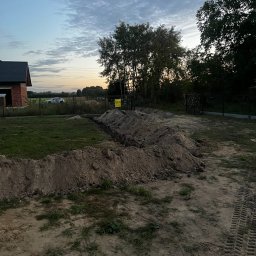 Michał - Wykopany rów w ziemi na działce, widoczne ślady opon, w tle dom w budowie i zachodzące słońce. Prace ziemne przygotowawcze do dalszych robót budowlanych.
