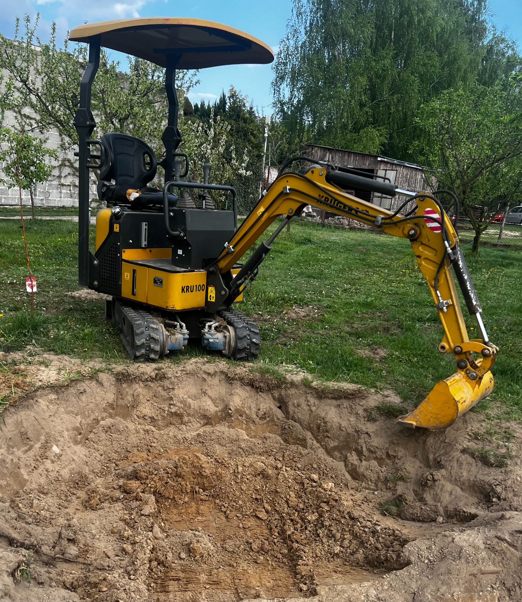 Żółta mini koparka KRU 100 w trakcie kopania okrągłego wykopu na trawiastym terenie, z budynkami i drzewami w tle.