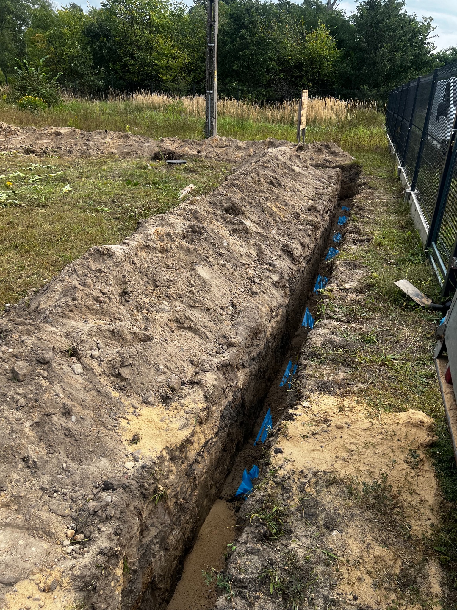 Wykopany rów z niebieskimi elementami w ziemi, widoczne przygotowania do instalacji. Rów otoczony ziemią i trawą, w tle drzewa i ogrodzenie.
