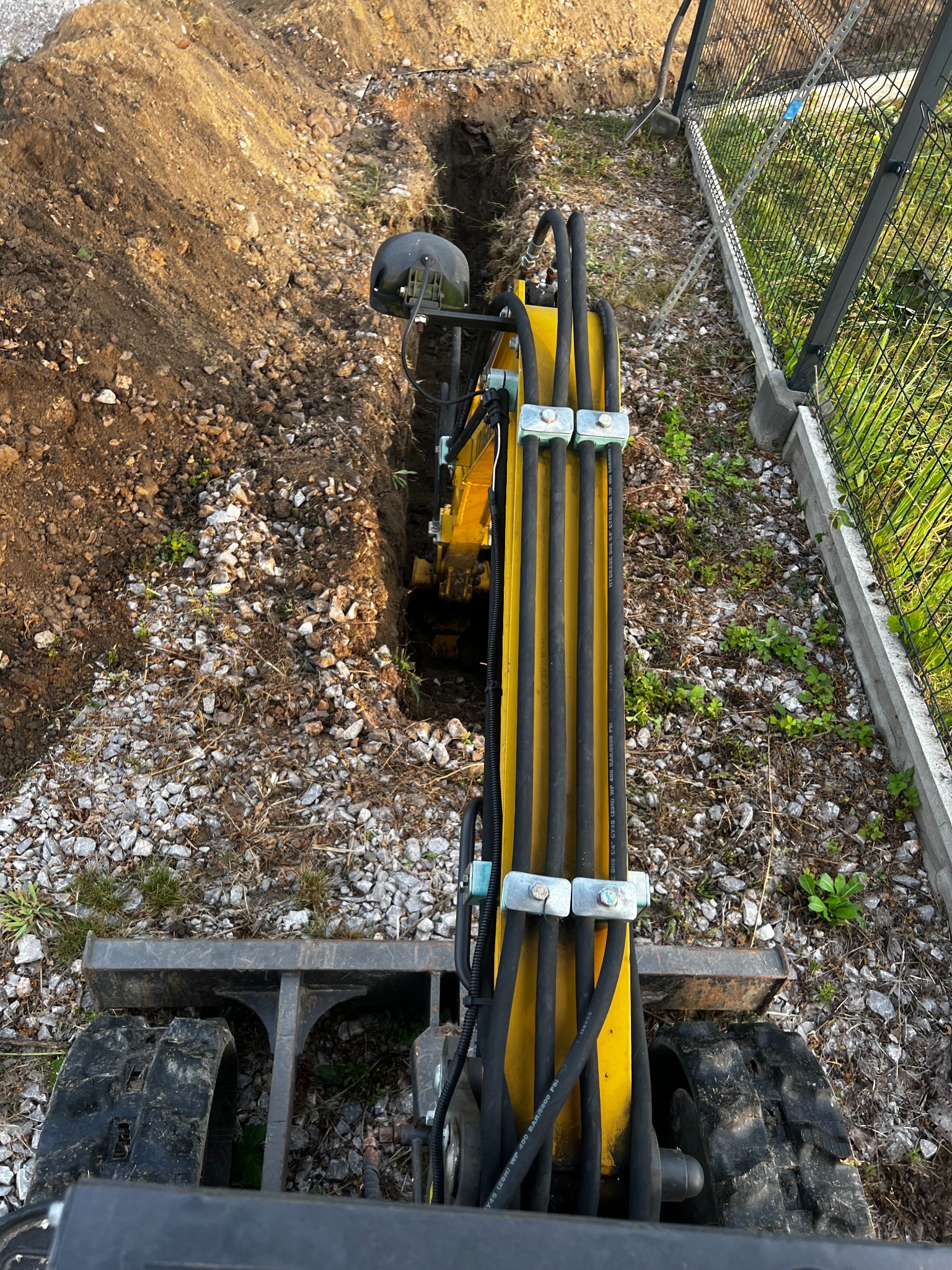Żółta minikoparka z widocznymi przewodami hydraulicznymi kopie wąski wykop w ziemi z kamieniami, obok ogrodzenie z siatki. Widok z góry na maszynę w trakcie pracy.