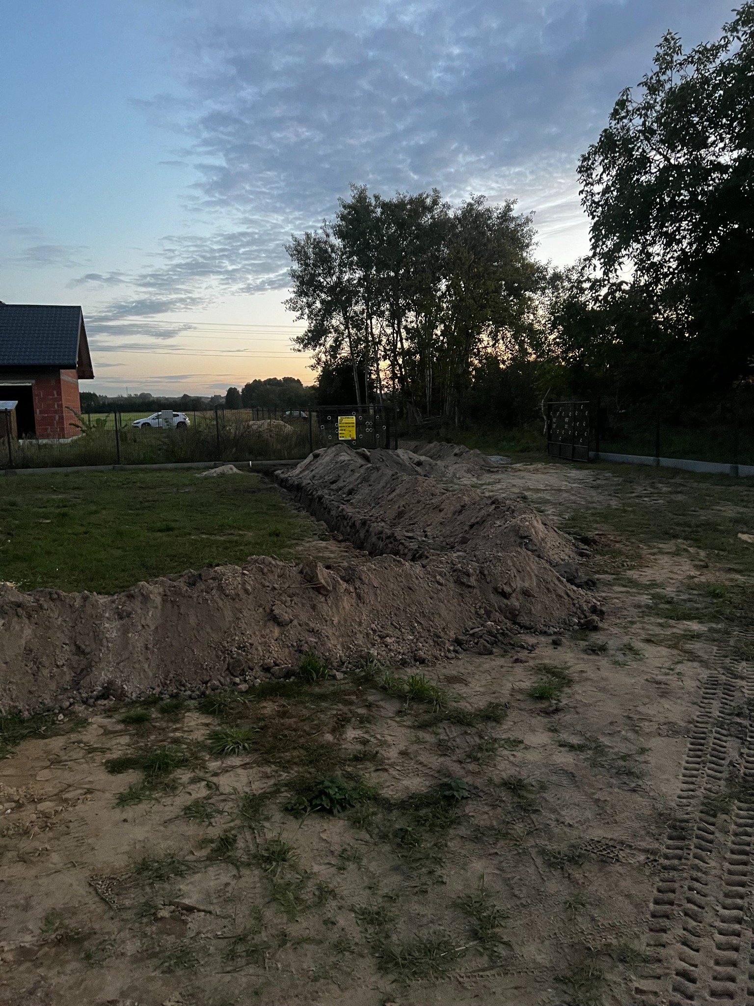 Wykopany rów w ziemi na działce, widoczne ślady opon, w tle dom w budowie i zachodzące słońce. Prace ziemne przygotowawcze do dalszych robót budowlanych.
