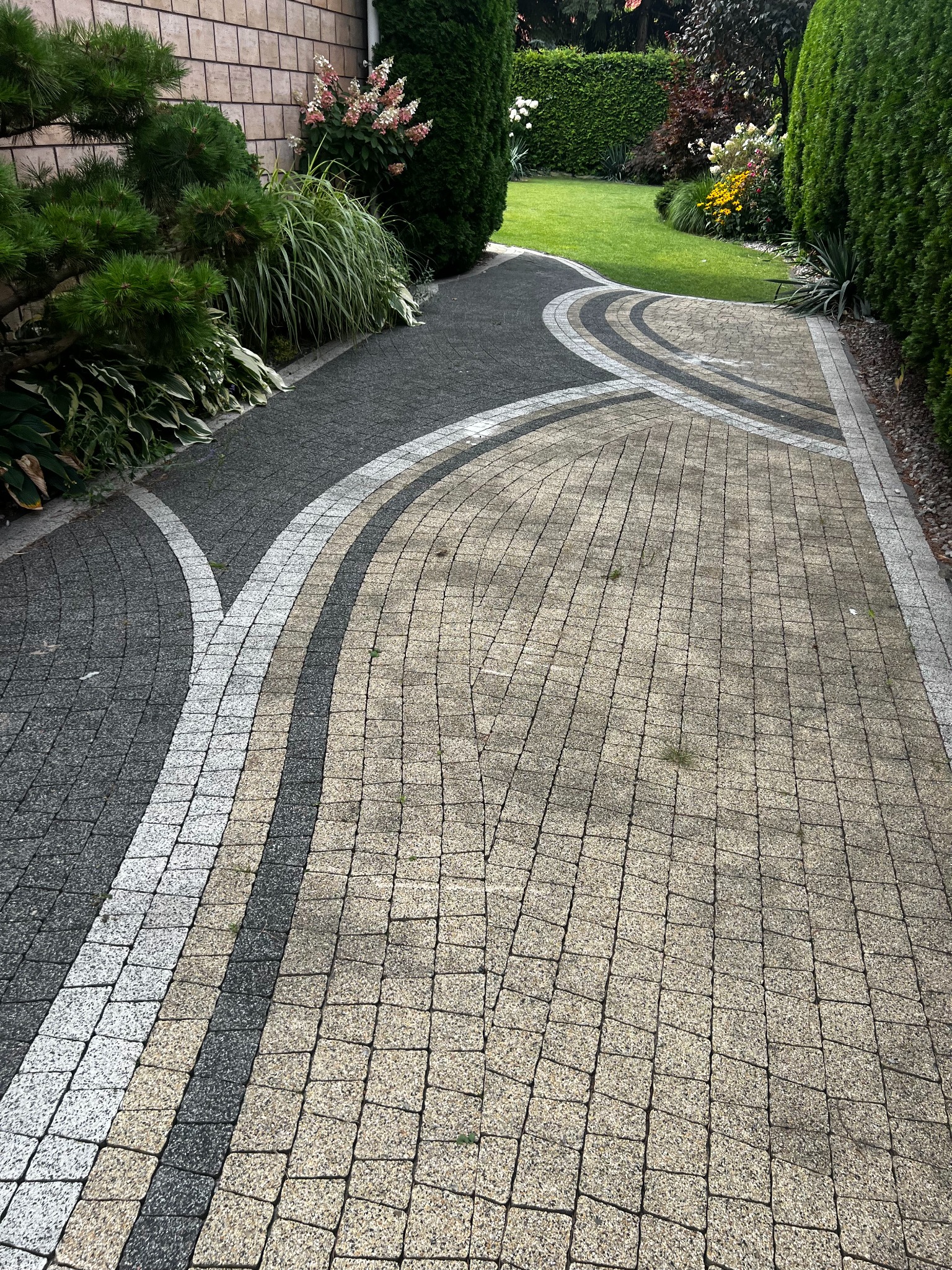 Kreatywny wzór ułożenia kostki brukowej na podjeździe, z wykorzystaniem odcieni szarości i beżu. W tle zadbany trawnik i roślinność.