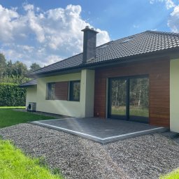 Radosław Poleszak - Parterowy dom z ciemną dachówką i drewnianą elewacją, otoczony zielonym trawnikiem i kamiennym podłożem. Widoczny komin i duże okna przesuwne.