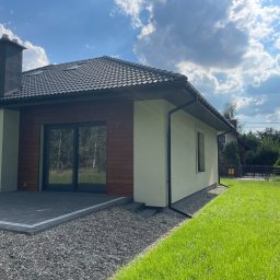 Radosław Poleszak - Gotowy, parterowy dom z ciemną dachówką i drewnianą elewacją, otoczony zielonym trawnikiem i szarą kostką brukową. Widok na fragment budynku w słoneczny dzień.