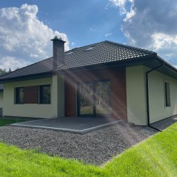 Radosław Poleszak - Parterowy dom z ciemną dachówką i drewnianą elewacją, otoczony zielonym trawnikiem i kamiennym podłożem. Widoczny komin i klimatyzator na ścianie. Słoneczny dzień.