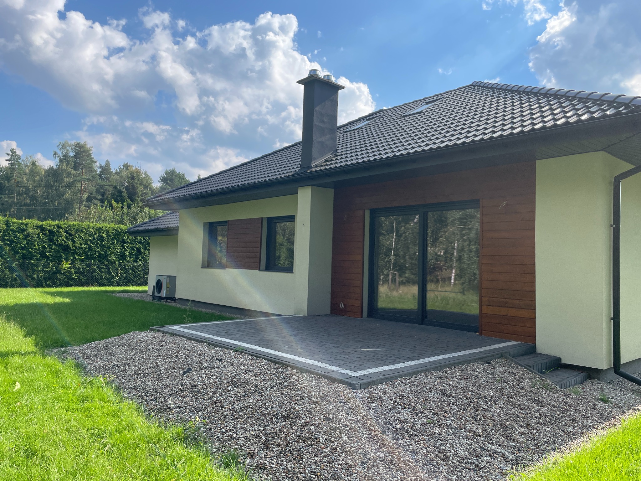 Parterowy dom z ciemną dachówką i drewnianą elewacją, otoczony zielonym trawnikiem i kamiennym podłożem. Widoczny komin i duże okna przesuwne.