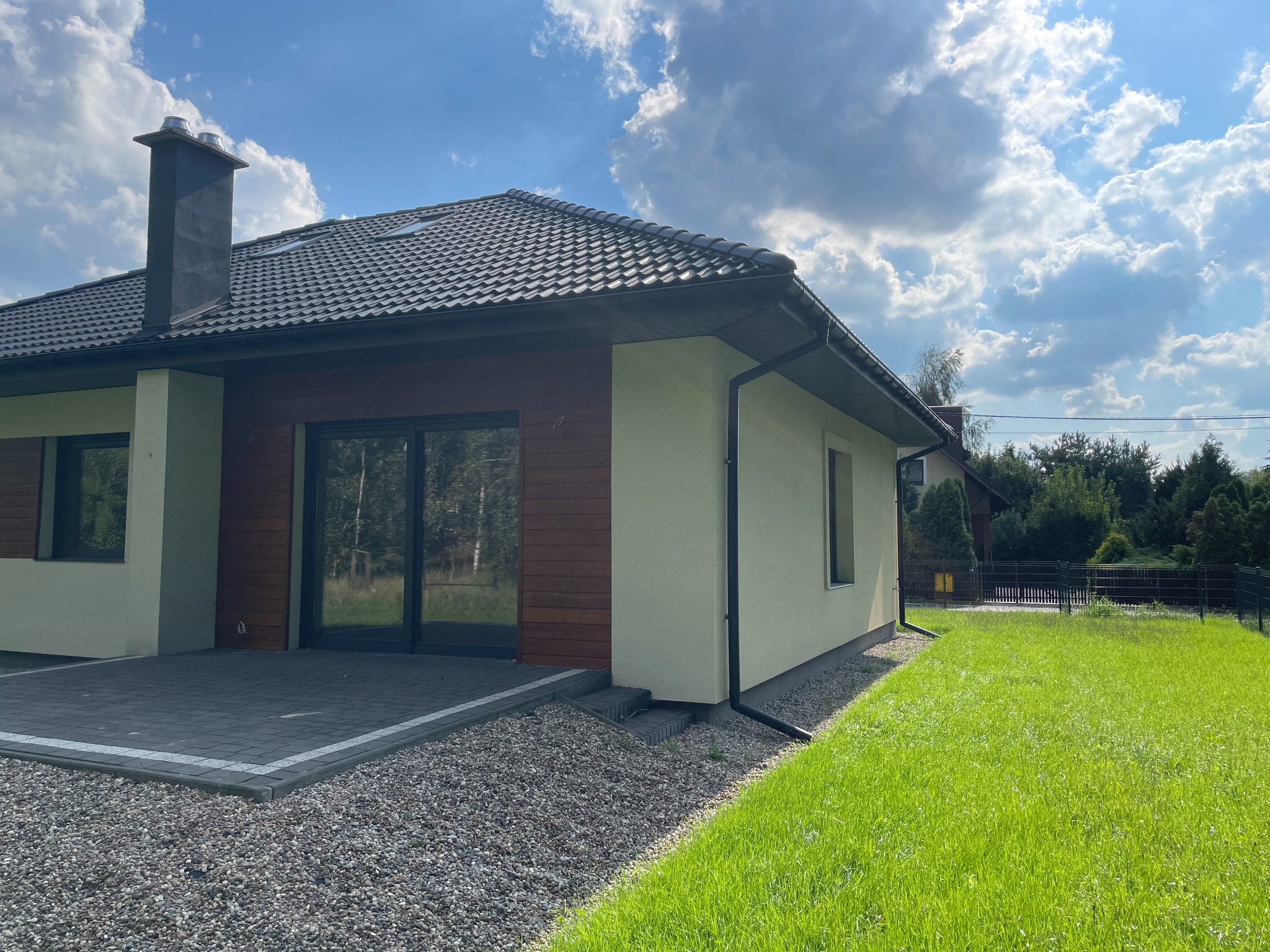 Gotowy, parterowy dom z ciemną dachówką i drewnianą elewacją, otoczony zielonym trawnikiem i szarą kostką brukową. Widok na fragment budynku w słoneczny dzień.