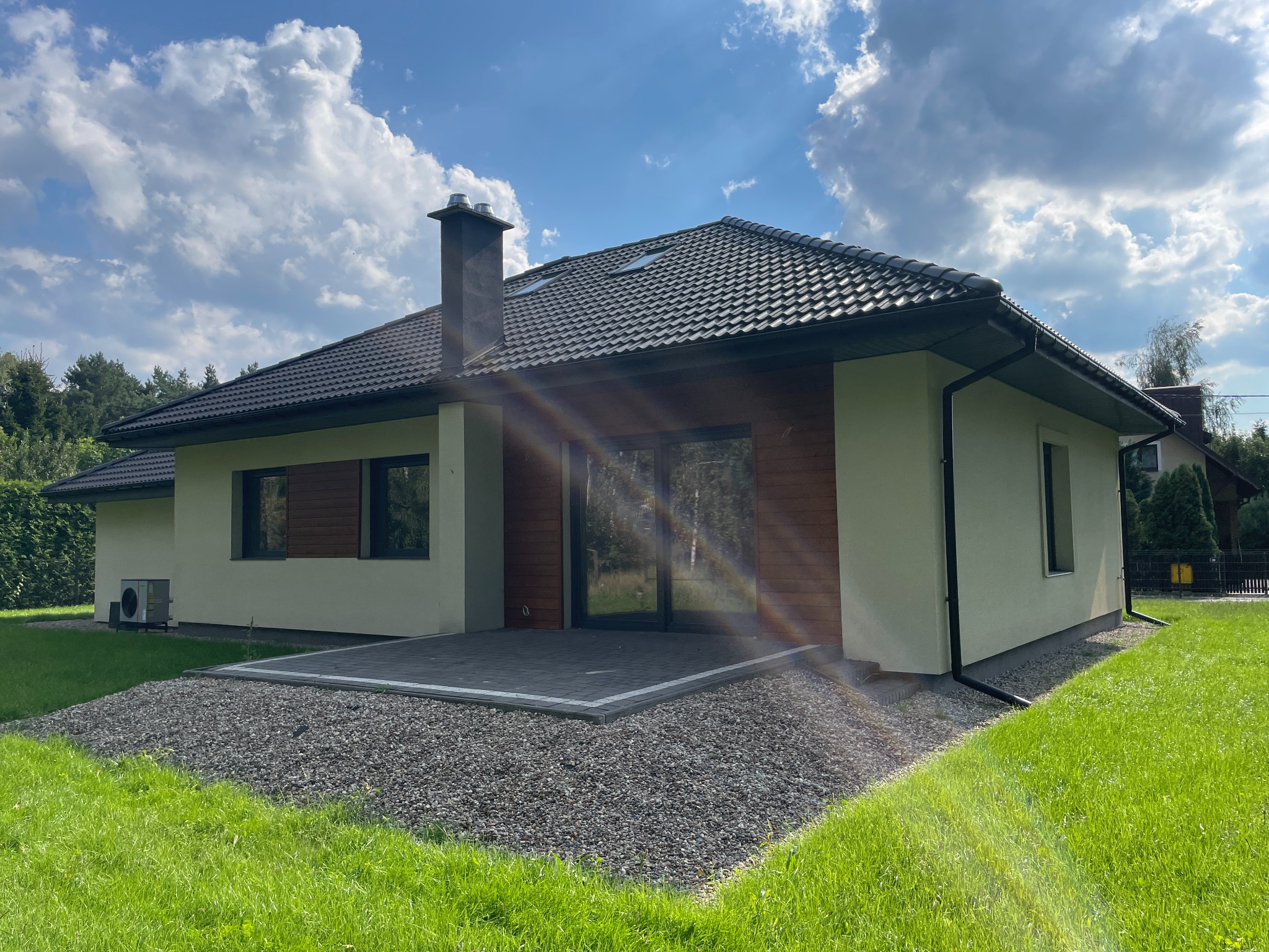 Parterowy dom z ciemną dachówką i drewnianą elewacją, otoczony zielonym trawnikiem i kamiennym podłożem. Widoczny komin i klimatyzator na ścianie. Słoneczny dzień.