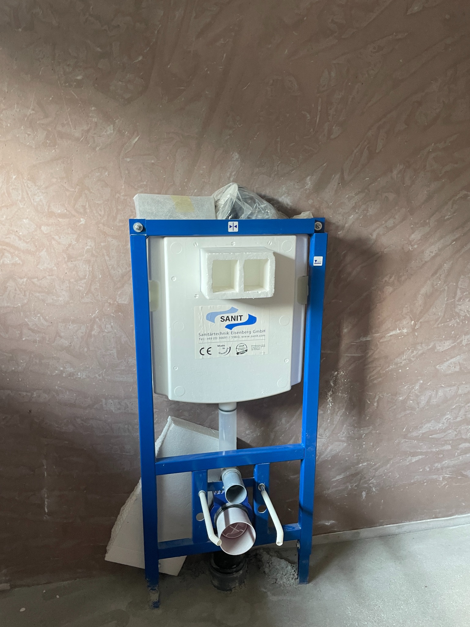 Stelaż podtynkowy SANIT w niebieskiej ramie, zamontowany przy surowej ścianie. Widoczne elementy instalacji wodno-kanalizacyjnej. Etap prac wykończeniowych w łazience.