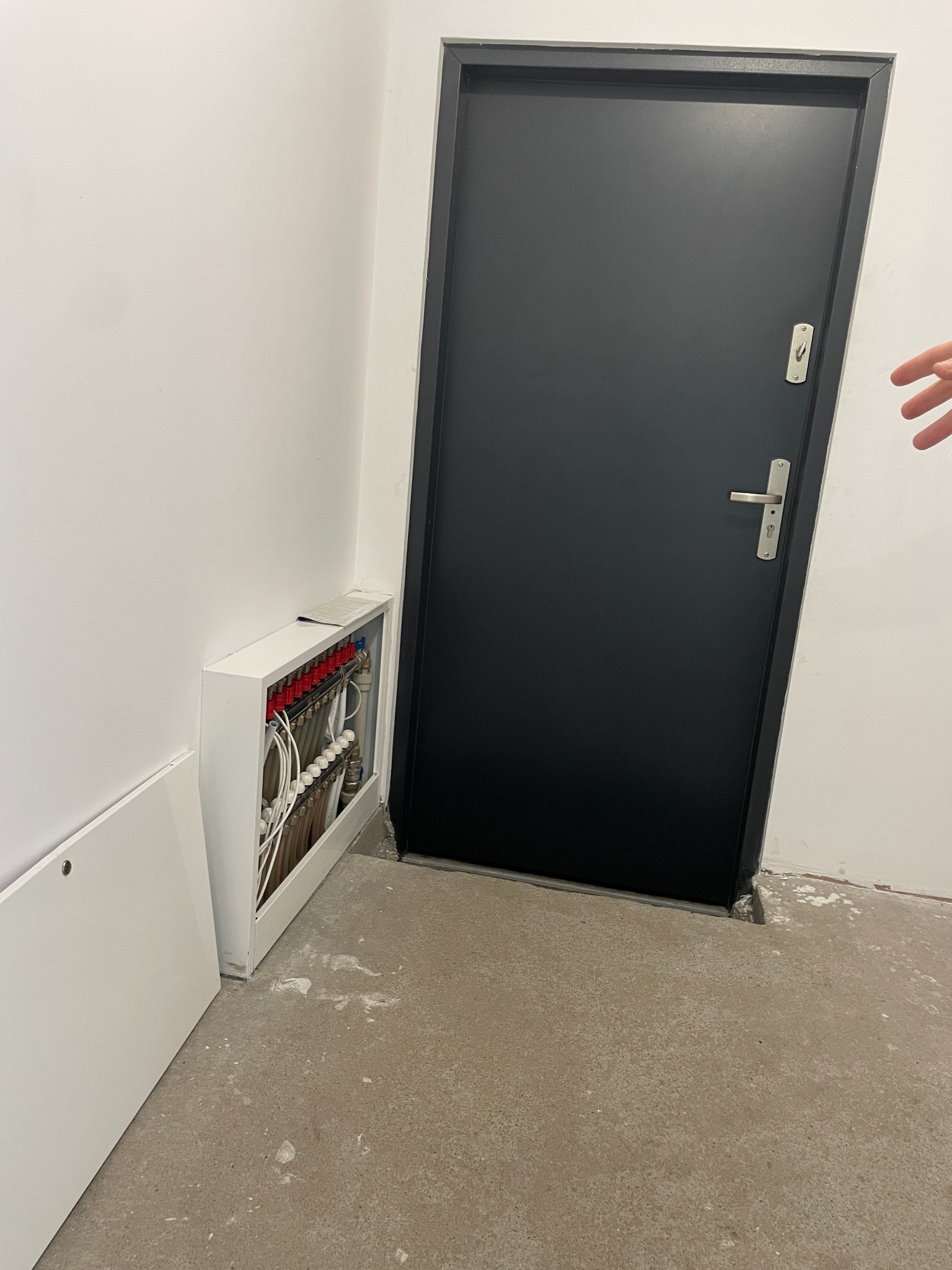 Rozdzielacz ogrzewania podłogowego w białej obudowie, zamontowany przy ścianie obok czarnych drzwi w surowym wnętrzu. Widoczna instalacja z rurkami i zaworami.
