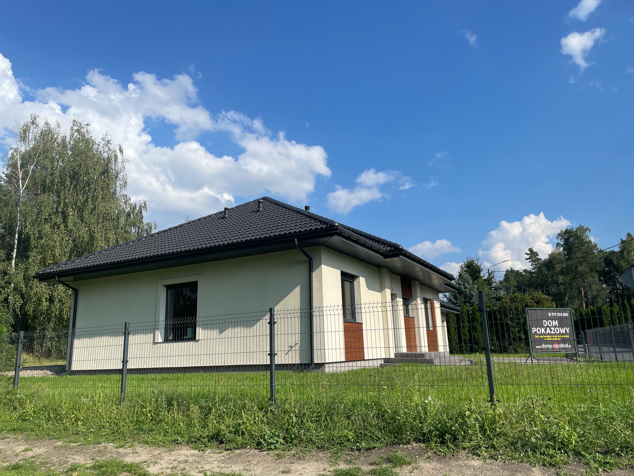 Gotowy dom pokazowy z ciemną dachówką i jasną elewacją, widoczny przez metalowe ogrodzenie. Na trawniku tablica informacyjna. Błękitne niebo z chmurami w tle.