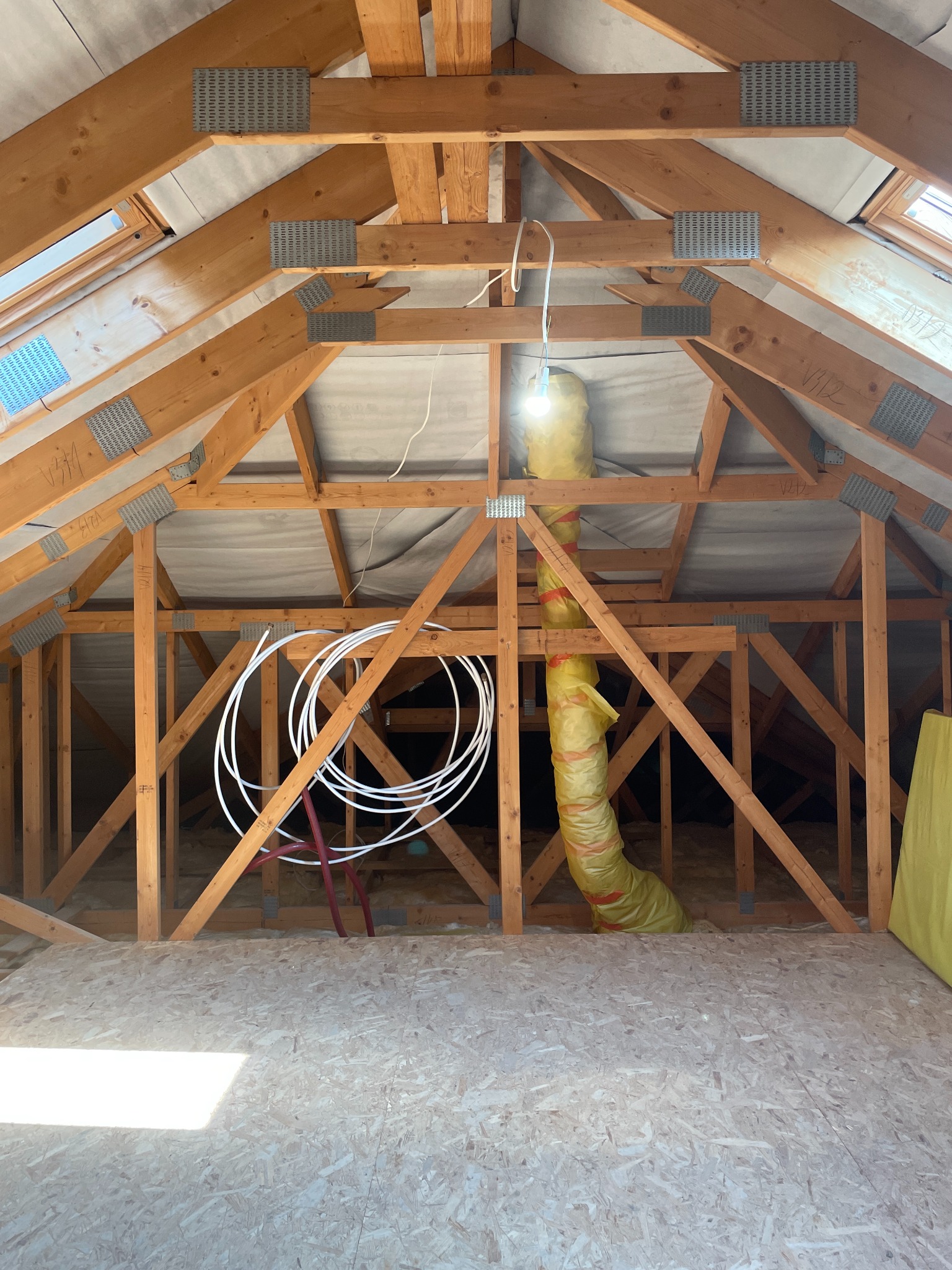 Wnętrze poddasza z drewnianą konstrukcją dachu, oknami połaciowymi i widoczną izolacją. Elementy instalacji wentylacyjnej i rury instalacyjne wewnątrz konstrukcji.