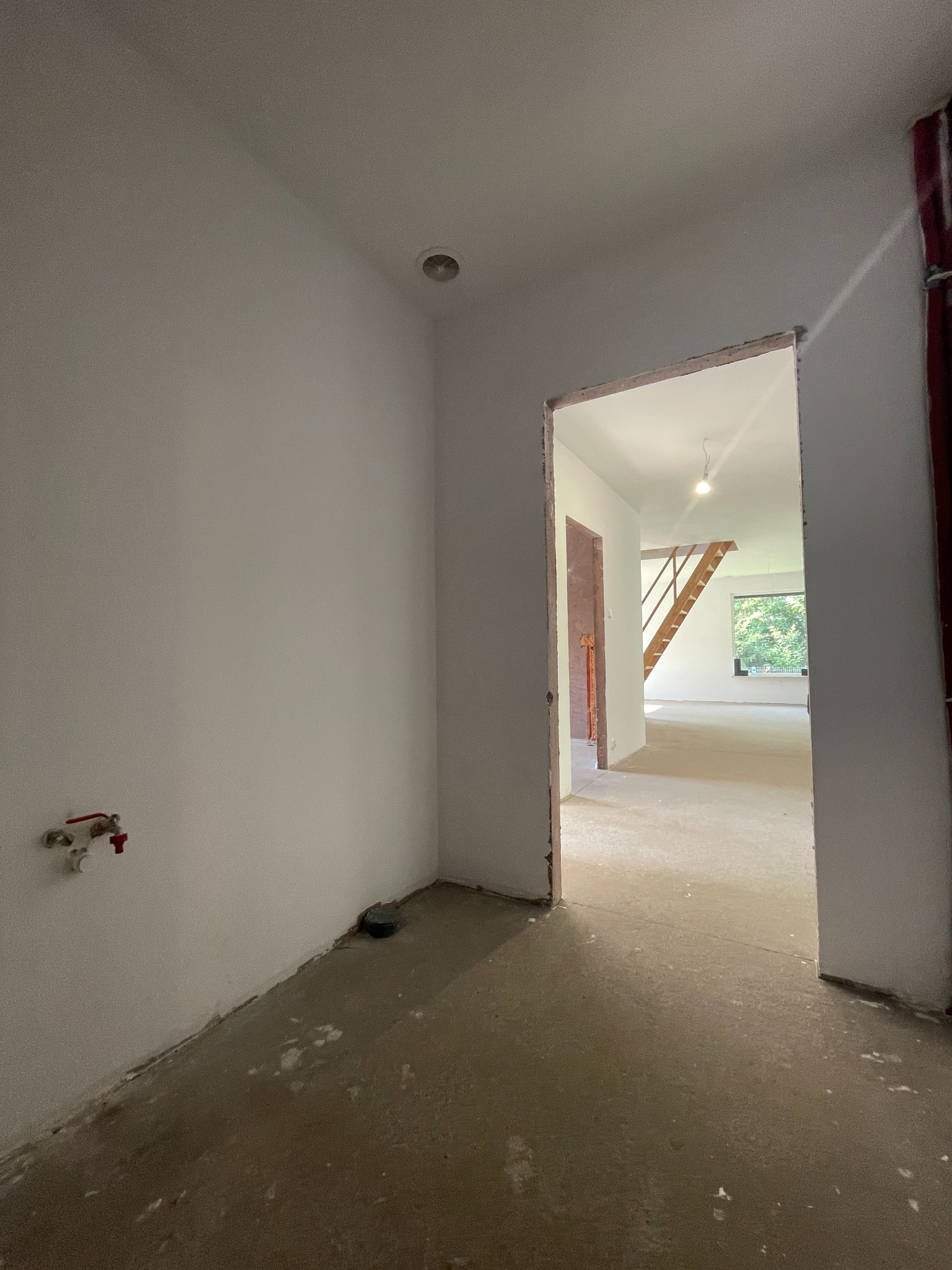 Wnętrze domu w stanie deweloperskim: surowe ściany, wylewka, widok na schody i okno przez otwarte drzwi. Instalacja wodna na ścianie.