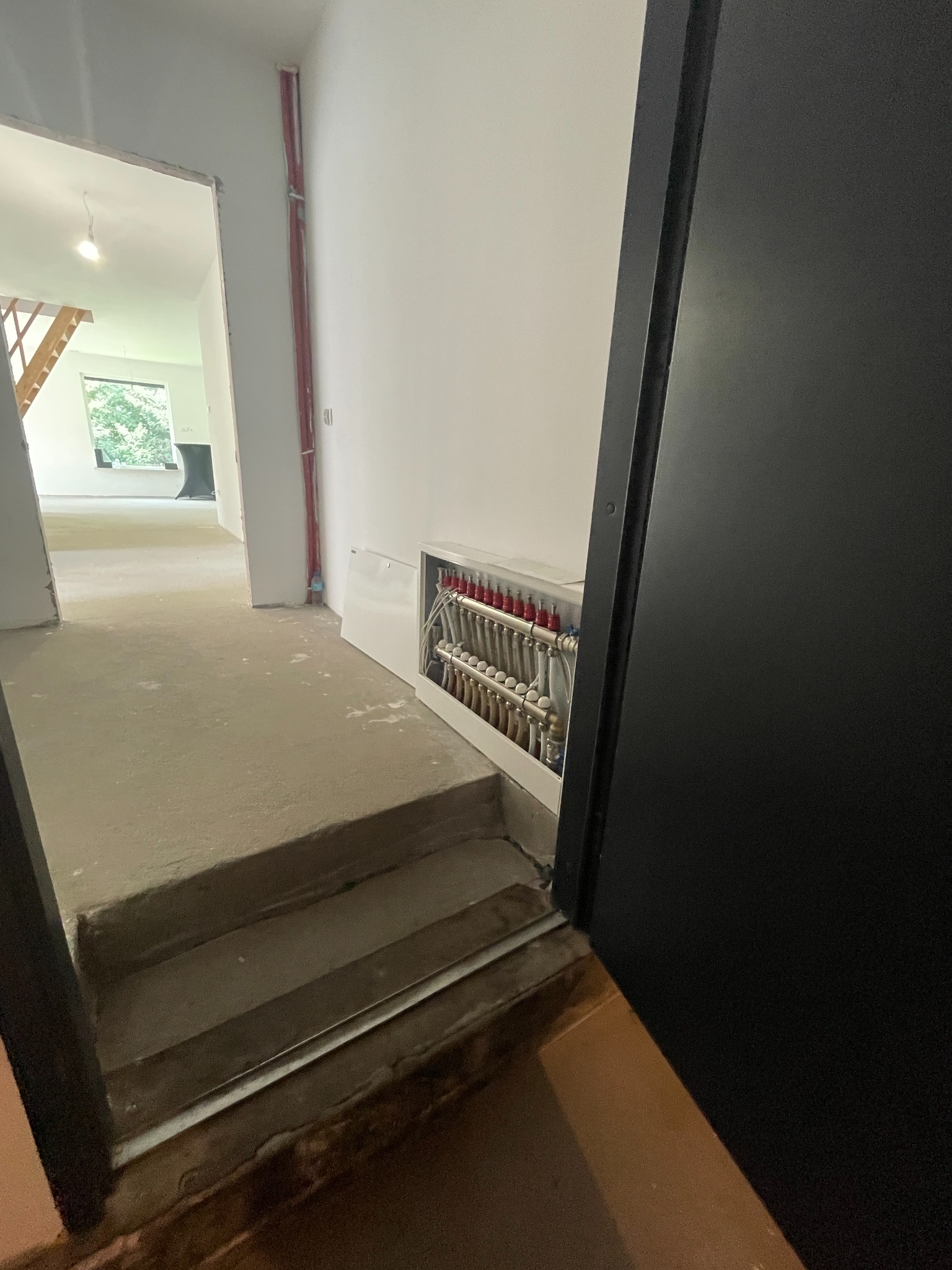 Wnętrze w trakcie budowy: widoczna instalacja ogrzewania podłogowego w rozdzielaczu, surowa wylewka i schody. W tle okno i drewniana konstrukcja schodów.