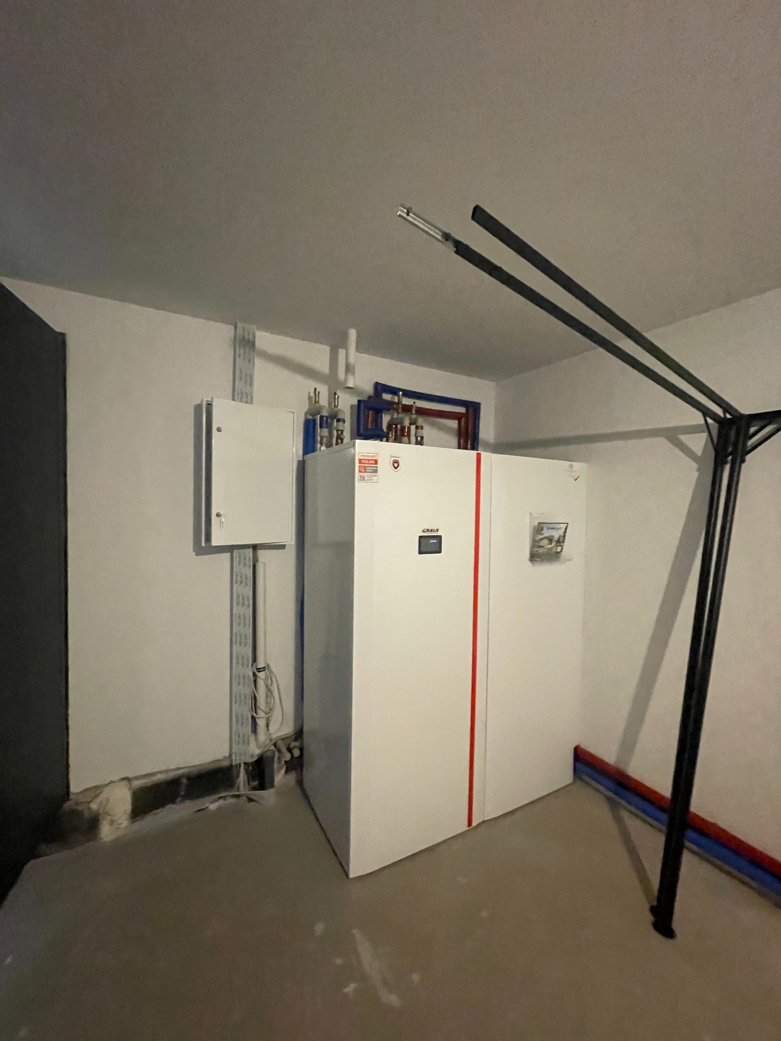 Biały piec grzewczy z czerwoną linią, zamontowany w pomieszczeniu z szarą podłogą i instalacjami na ścianie. Widoczne rury i skrzynka elektryczna.