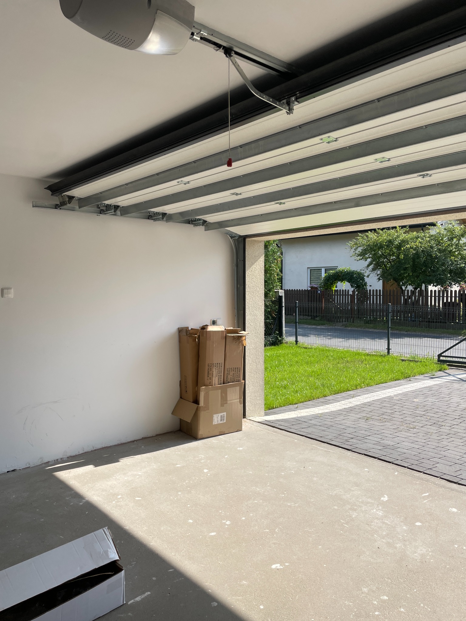 Wnętrze garażu w stanie surowym z otwartą bramą segmentową, widok na posesję z kostką brukową i zielenią. Na podłodze leżą kartony, sufit z widocznym mechanizmem bramy.