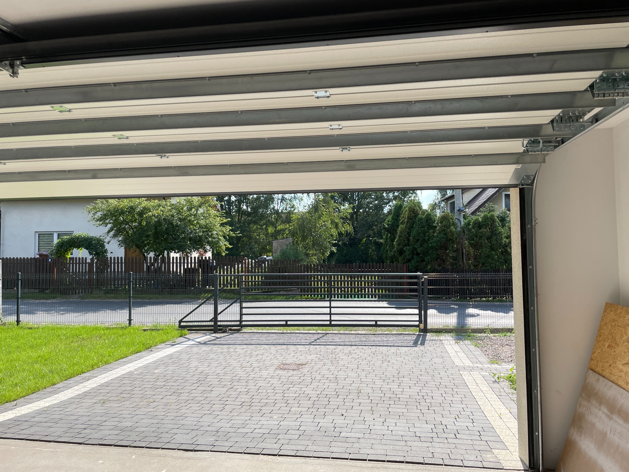Otwarta brama garażowa z widokiem na podjazd z kostki, ogrodzenie i zieleń. Jasne, słoneczne oświetlenie. Widok z wnętrza garażu na zewnątrz.