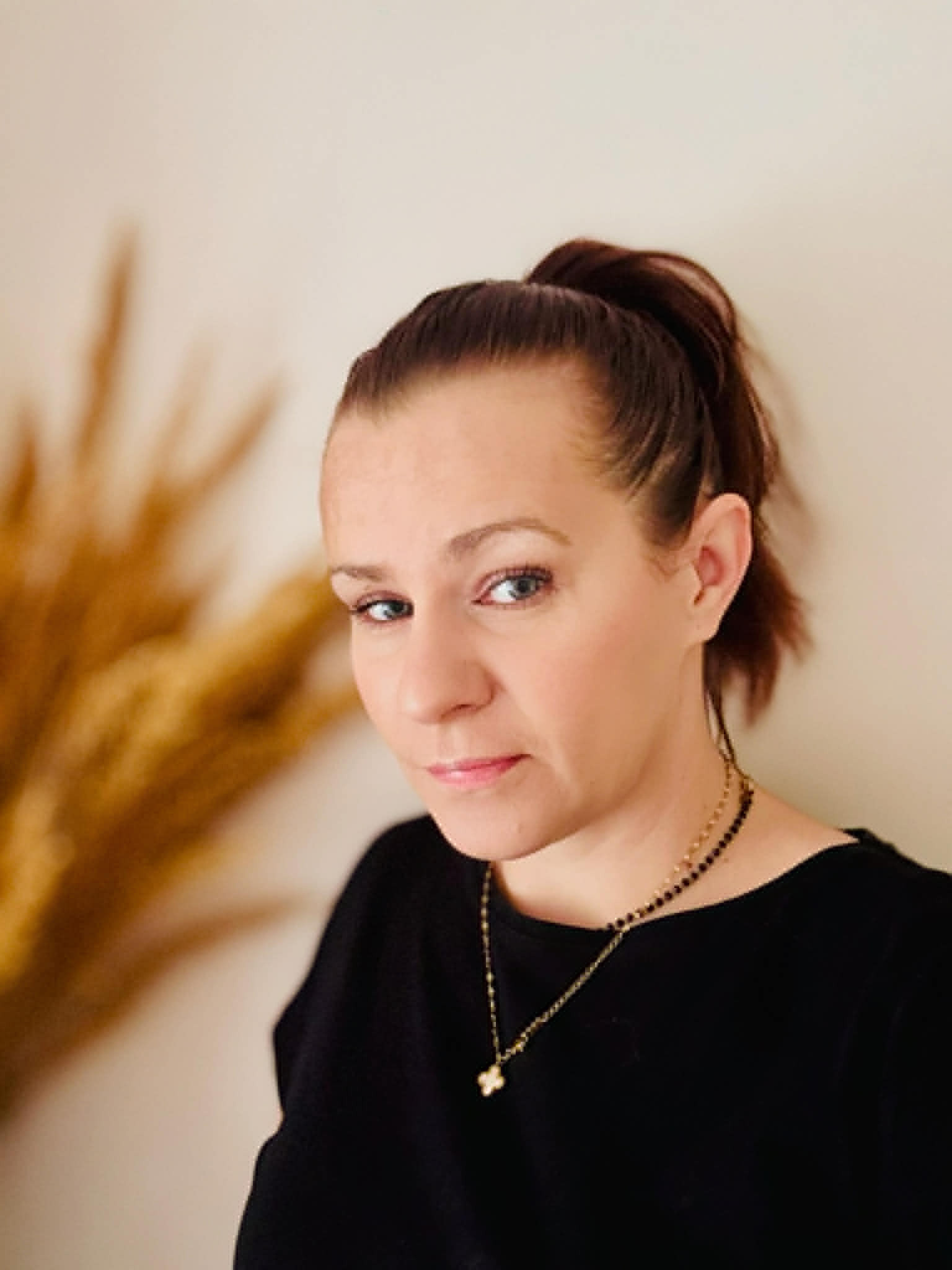 Portret kobiety z brązowymi włosami spiętymi w kucyk, w czarnej bluzce i ze złotym naszyjnikiem, na rozmytym tle z dekoracyjną trawą.