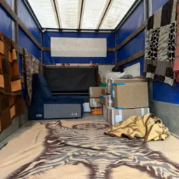 Wnętrze busa transportowego z zapakowanymi kartonami, fotelem i pledami. Widoczne niebieskie ściany i biały dach pojazdu. Perspektywa od tyłu pojazdu.