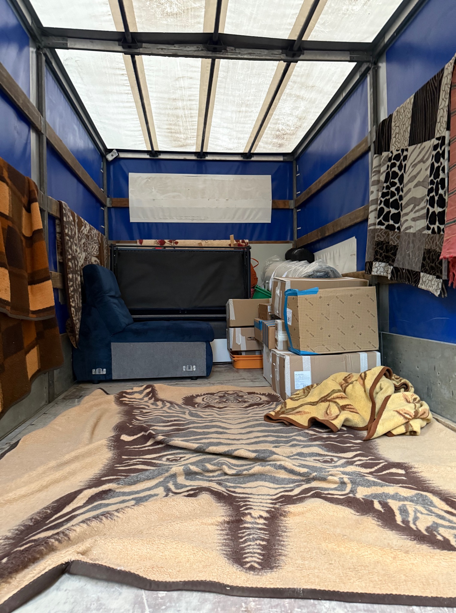 Wnętrze busa transportowego z zapakowanymi kartonami, fotelem i pledami. Widoczne niebieskie ściany i biały dach pojazdu. Perspektywa od tyłu pojazdu.