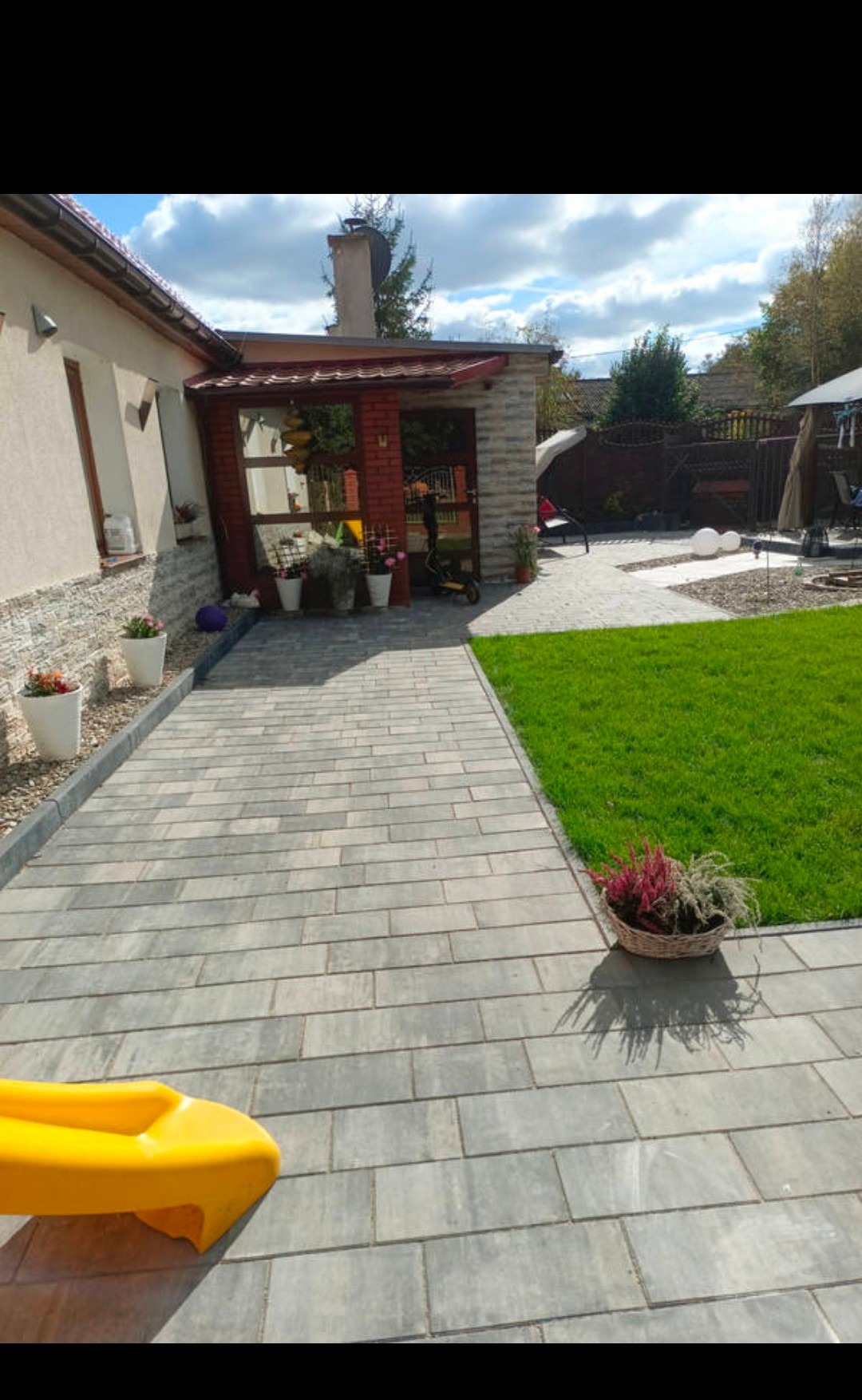 Ułożona szara kostka brukowa prowadzi od domu do ogrodu. W tle widoczny budynek z ceglanym gankiem i zielony trawnik. Wzdłuż ścieżki donice z kwiatami.