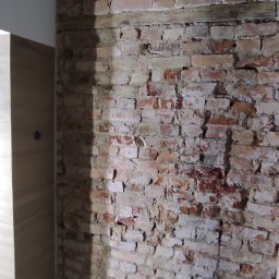 UJUT GROUP SPÓŁKA Z OGRANICZONĄ ODPOWIEDZIALNOŚCIĄ - Fragment wnętrza z odsłoniętą ceglaną ścianą z łukowym nadprożem, drewnianą belką i wzorzystą podłogą. Widoczny fragment szafki.