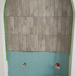 UJUT GROUP SPÓŁKA Z OGRANICZONĄ ODPOWIEDZIALNOŚCIĄ - Wnęka z wykończonym fragmentem ściany płytkami w odcieniach szarości, przygotowana do dalszych prac wykończeniowych. Widoczne kartony i narzędzia.