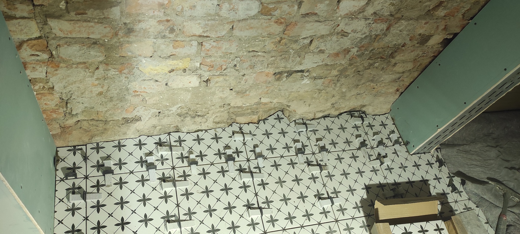 Układanie płytek z wzorem kwiatowym na podłodze łazienki, widoczne krzyżyki dystansowe i fragment surowego muru oraz płyta gipsowa w rogu.