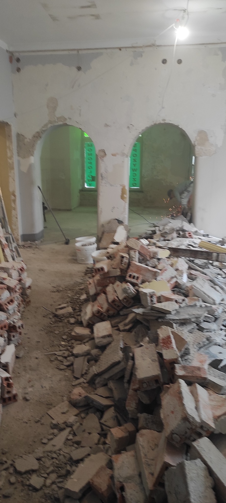 Wnętrze w trakcie rozbiórki: sterta gruzu ceglanego, widoczne łuki w ścianach, zabezpieczone okna folią. Prace remontowe w toku, widoczne iskry od szlifierki w tle.