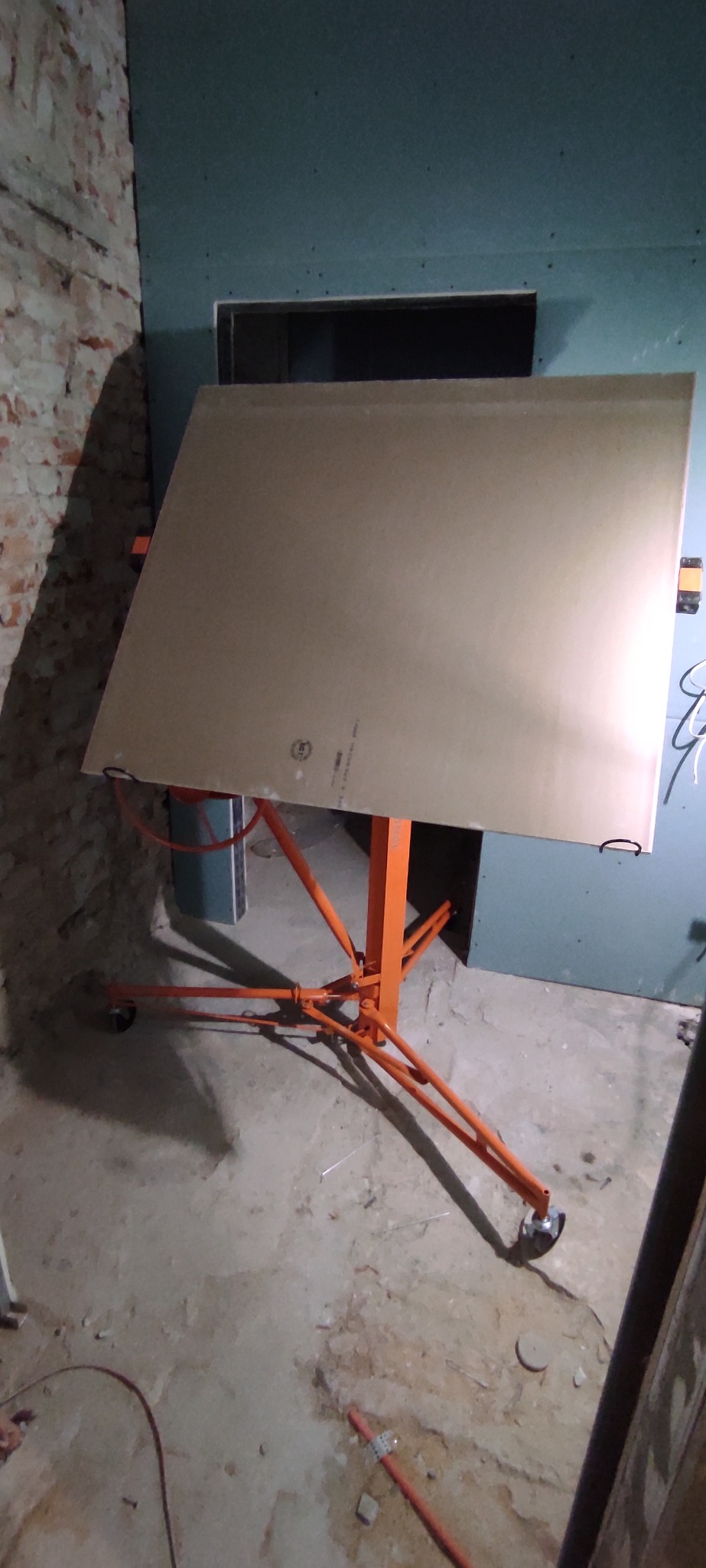 Płyta gipsowo-kartonowa na pomarańczowym stelażu z kółkami, przygotowana do montażu na ścianie z płyt gipsowych. Widoczny fragment ściany z cegły.