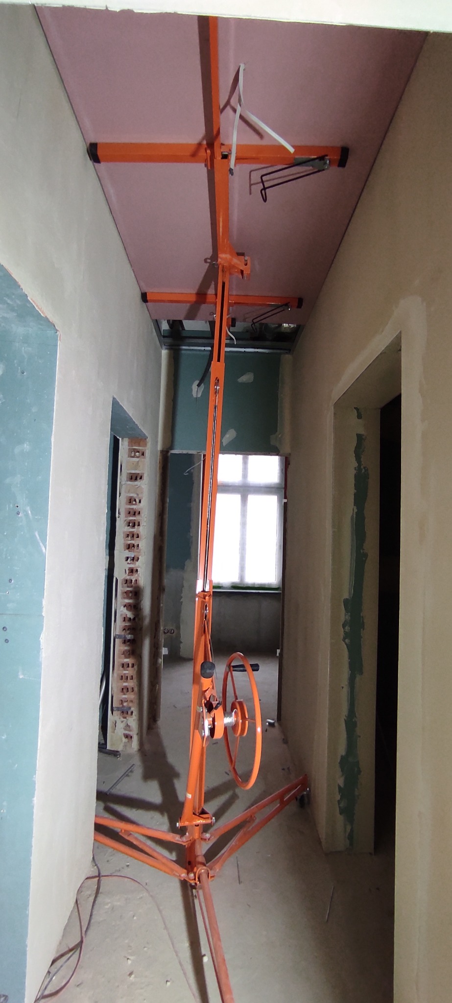 Pomarańczowy podnośnik do płyt gipsowych montuje różową płytę na suficie w remontowanym pomieszczeniu. Widoczne ściany w trakcie szpachlowania i okno w tle.