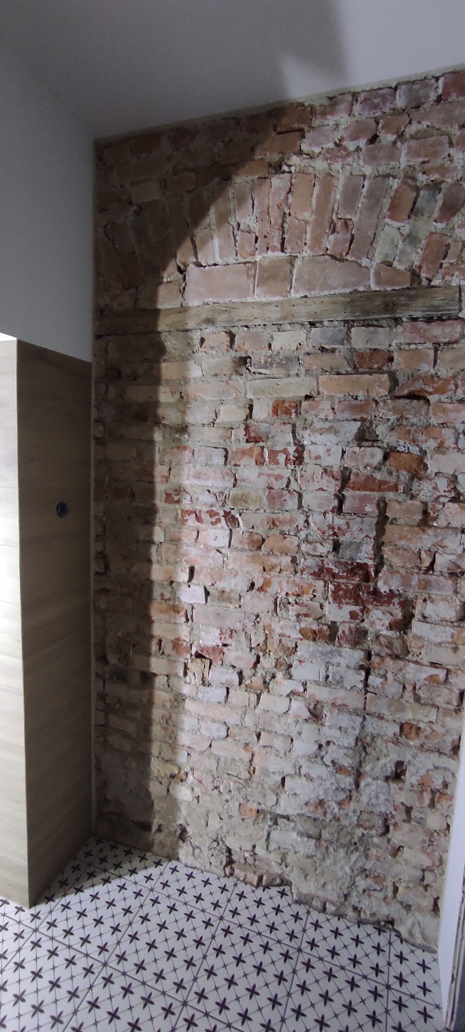 Fragment wnętrza z odsłoniętą ceglaną ścianą z łukowym nadprożem, drewnianą belką i wzorzystą podłogą. Widoczny fragment szafki.