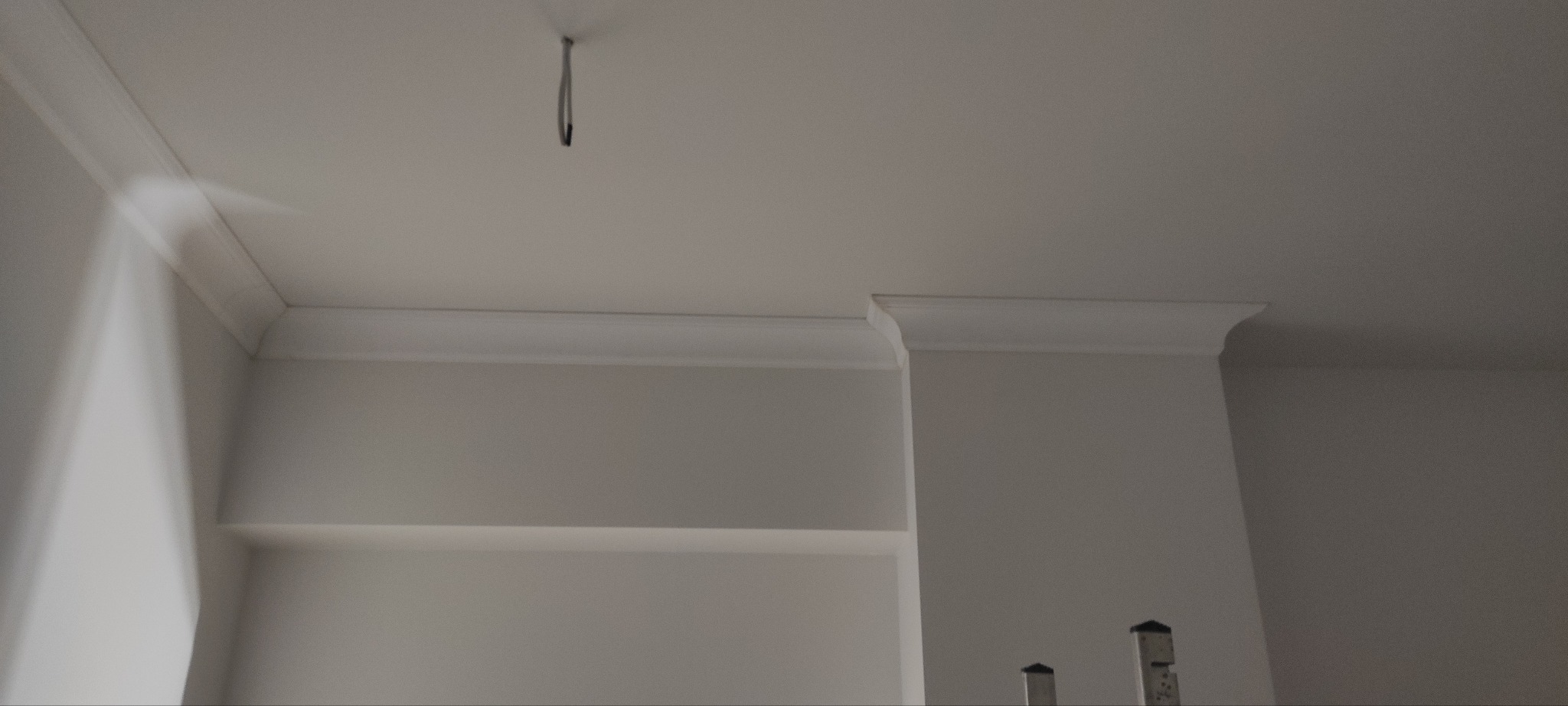 Wykończony fragment wnętrza z listwami dekoracyjnymi przy suficie i półkami. Widoczny kabel ze ściany. Całość w jasnej kolorystyce.