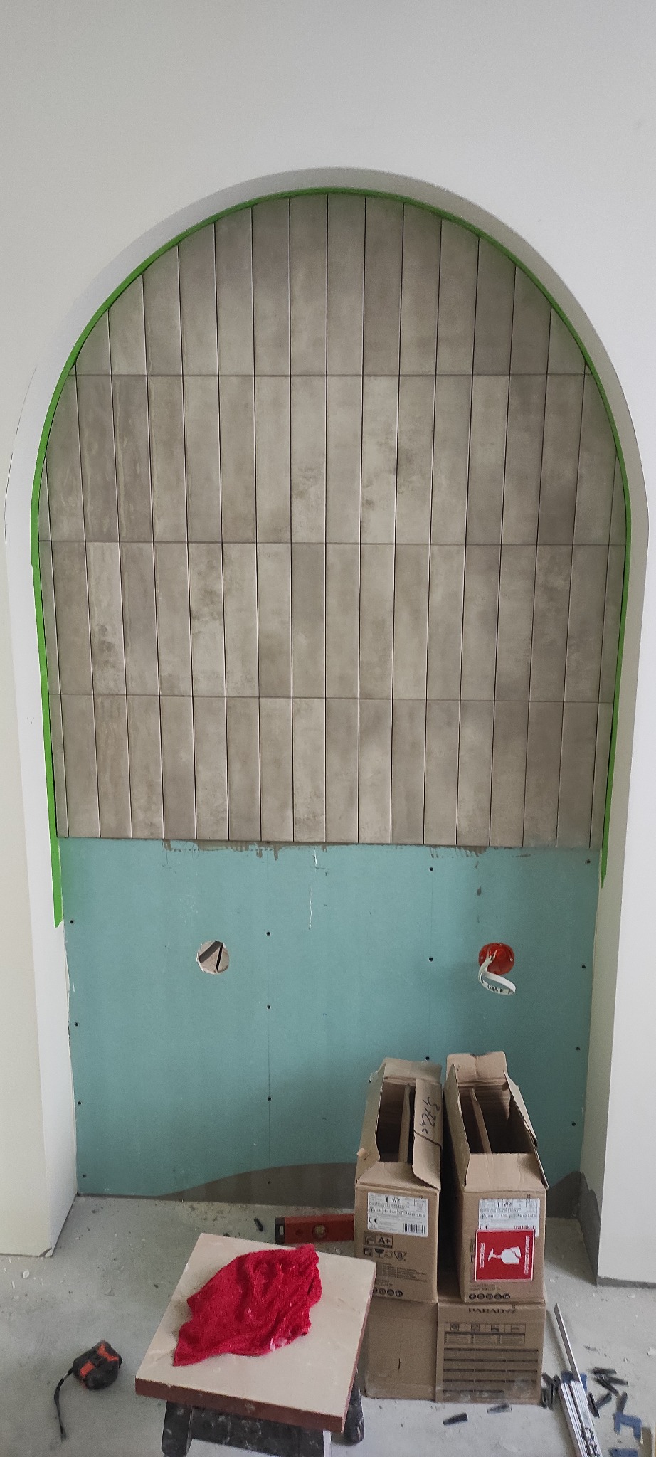 Wnęka z wykończonym fragmentem ściany płytkami w odcieniach szarości, przygotowana do dalszych prac wykończeniowych. Widoczne kartony i narzędzia.