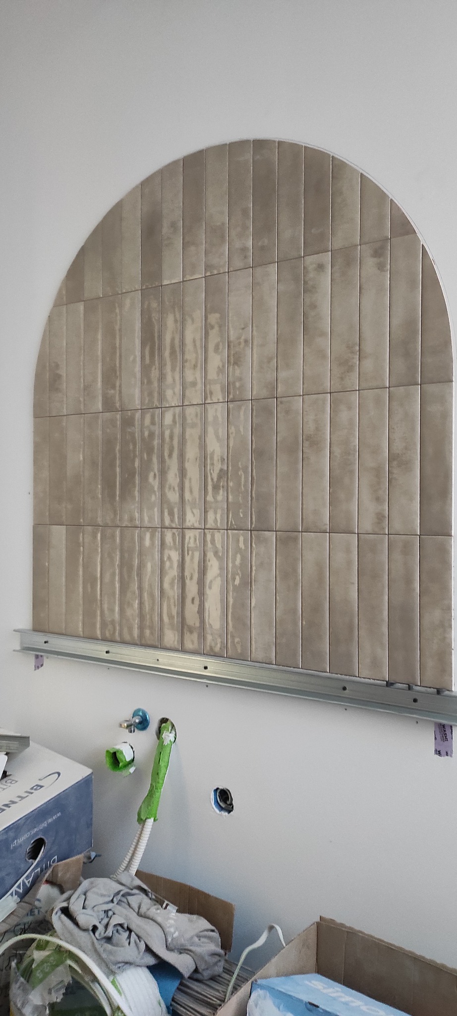 Aranżacja łazienki z półokrągłą ścianką wyłożoną beżowymi, pionowymi płytkami na metalowej konstrukcji. Widoczne przyłącza wodne i kartony po płytkach.