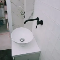 Biała, okrągła umywalka nablatowa z czarną armaturą na szafce w łazience z białymi płytkami i marmurowym akcentem. Widoczne przewody elektryczne.