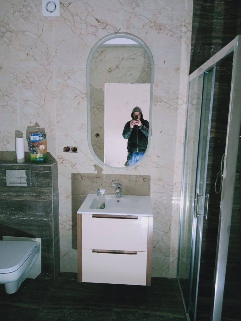 Wykończona łazienka z kabiną prysznicową, umywalką z szafką i owalnym lustrem, odbijającym osobę robiącą zdjęcie telefonem. Marmurowe płytki na ścianach i podłodze.