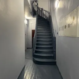 Klatka schodowa po malowaniu: szare ściany, drewniane schody i podłoga w odcieniach szarości. Widoczne skrzynki na listy i oświetlenie. Ujęcie z dołu schodów.