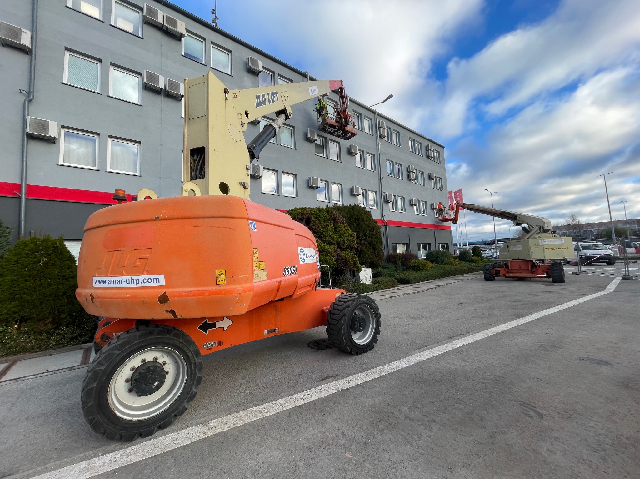 Dwie pomarańczowe platformy podnośnikowe JLG Lift na tle szarego budynku z klimatyzacją i błękitnego nieba. Pracownicy w koszach platform wykonują prace na wysokości.