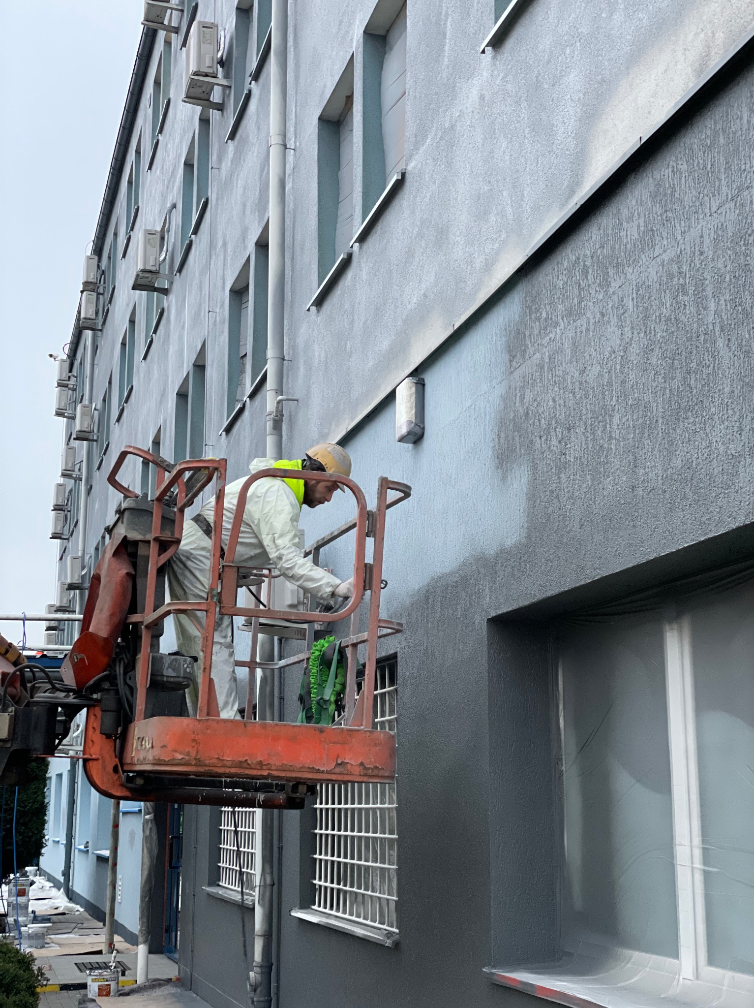 Pracownik w kombinezonie maluje elewację budynku z podnośnika koszowego na szaro. Okna zabezpieczone folią. Widoczne klimatyzatory na ścianie.