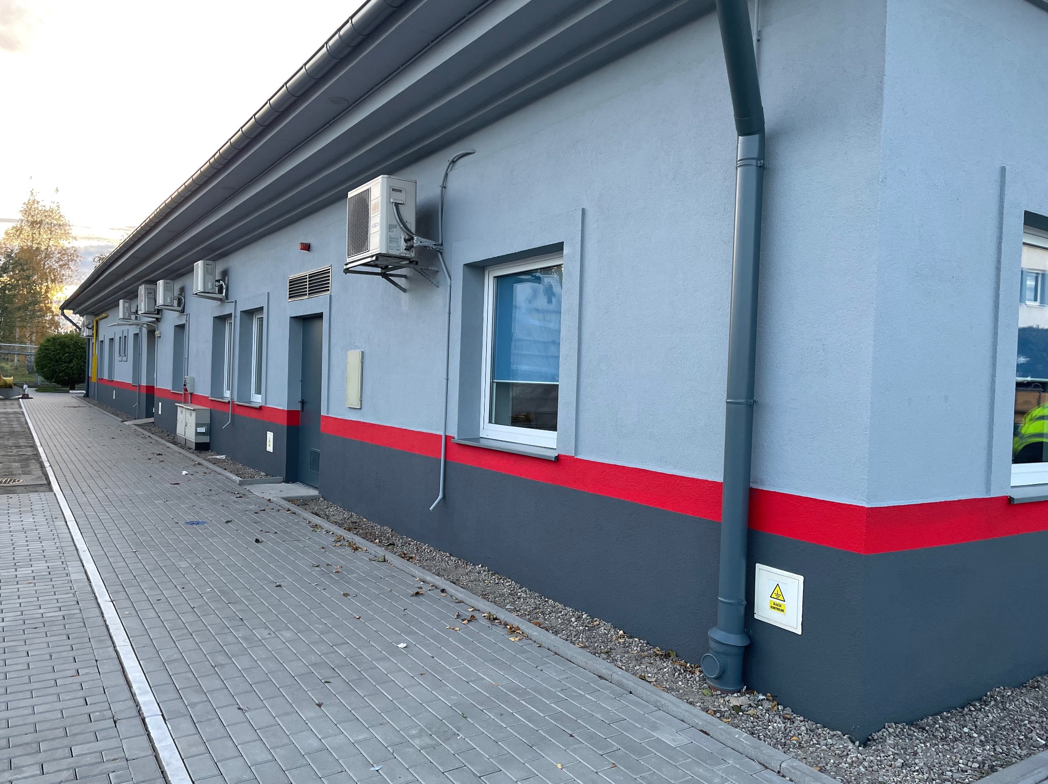 Odnowiona elewacja budynku z szarą farbą, czerwoną linią i ciemnoszarym cokołem. Widoczne okna, klimatyzatory i rynna. Kostka brukowa przed budynkiem.
