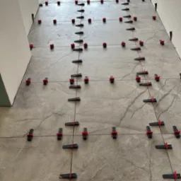 Szare płytki podłogowe z marmurkowym wzorem, wyrównane systemem poziomowania płytek z czarnymi klinami i czerwonymi klipsami, w trakcie układania na korytarzu.