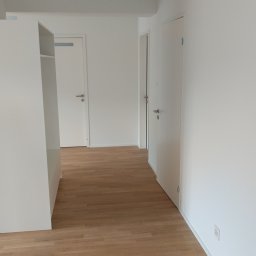 Daniel Sawiak - Jasne, odnowione wnętrze z drewnianą podłogą i białymi ścianami. Widoczne drzwi i fragment białej szafy. Minimalistyczny styl, świeżo po remoncie.