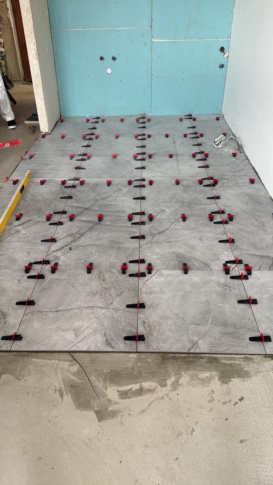 Szare, marmurkowe płytki podłogowe w trakcie układania, z czerwonymi klipsami poziomującymi i niebieską płytą kartonowo-gipsową w tle. Widoczny poziomica.