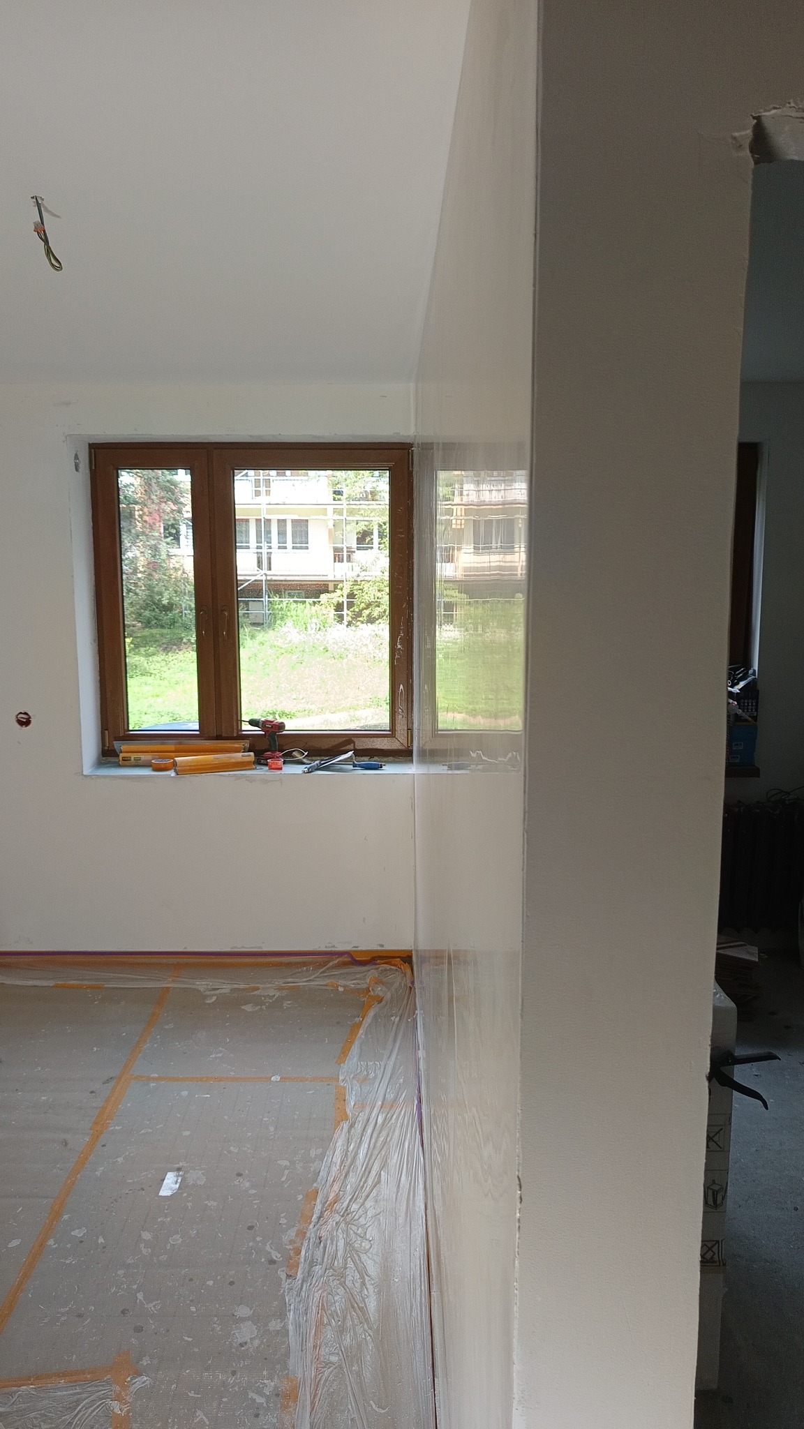 Pokój w trakcie prac wykończeniowych: zabezpieczona podłoga folią, ściany po szpachlowaniu, narzędzia na parapecie drewnianego okna.