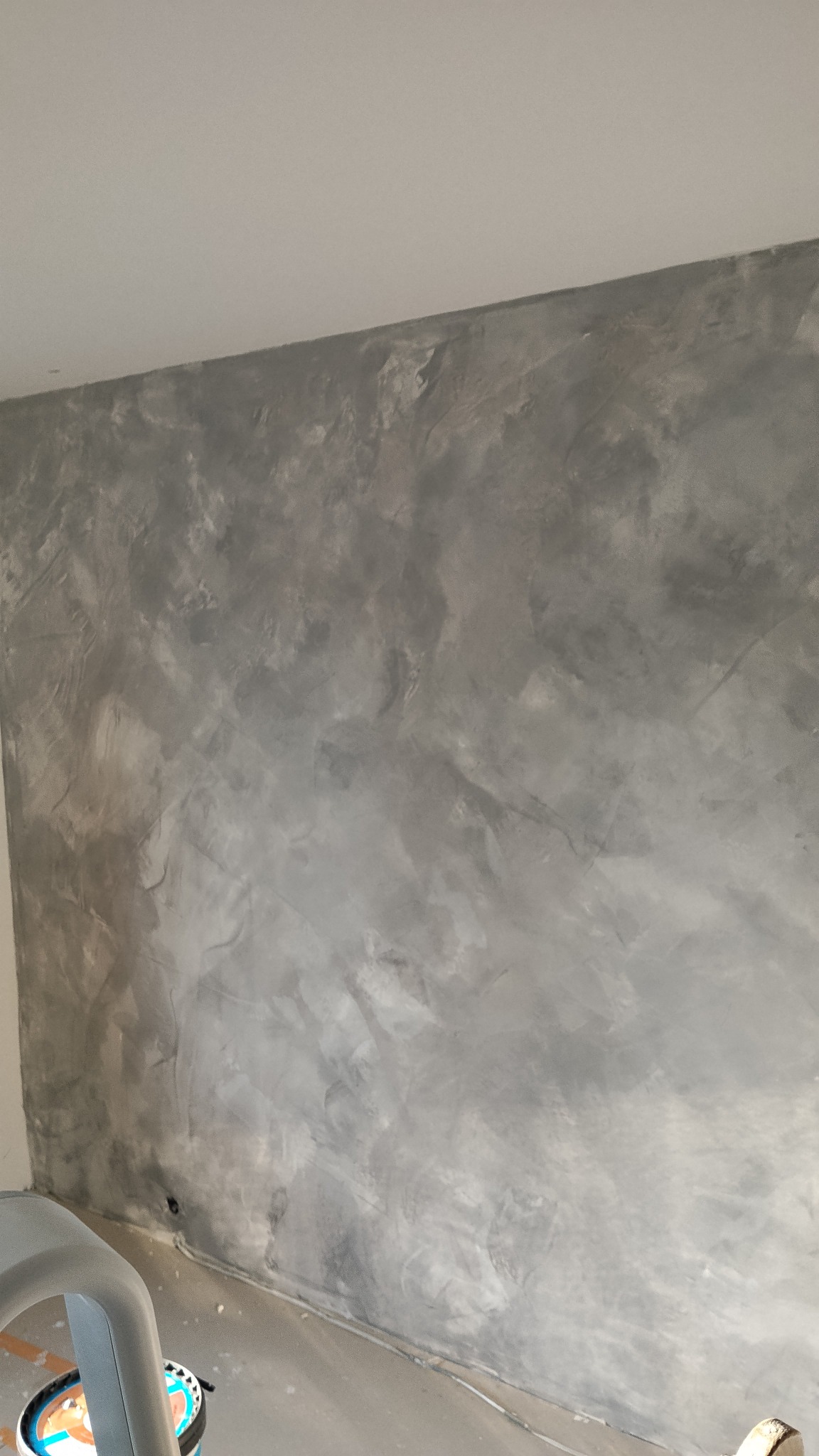 Szara, dekoracyjna ściana w stylu betonu architektonicznego, z widocznymi niuansami kolorystycznymi i fakturą. U dołu fragment podłogi i narożnik ściany.