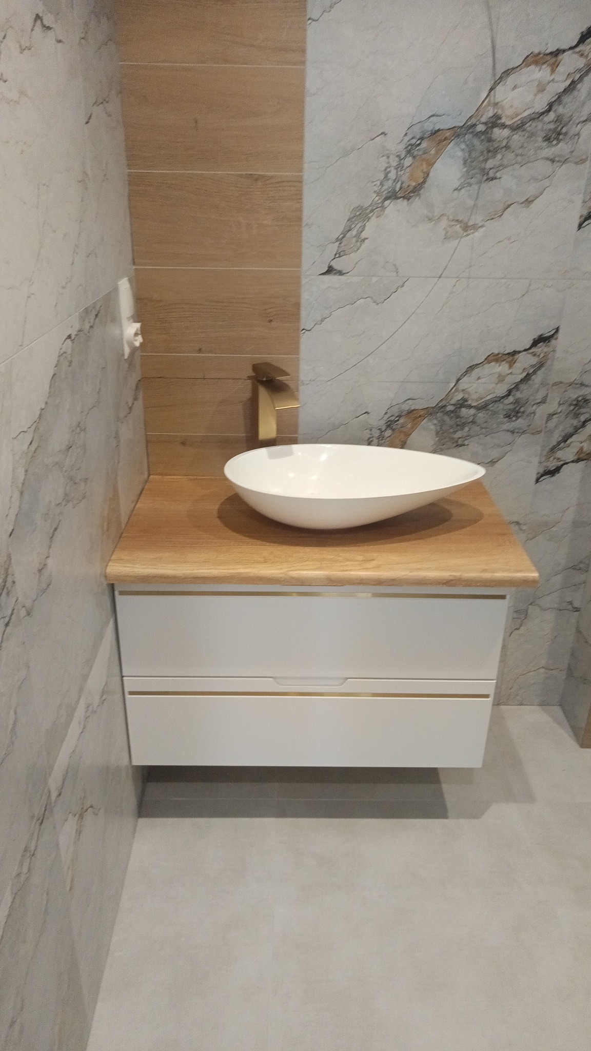 Nowoczesna łazienka: biała szafka z umywalką nablatową i złotą armaturą na tle płytek imitujących marmur i drewno.